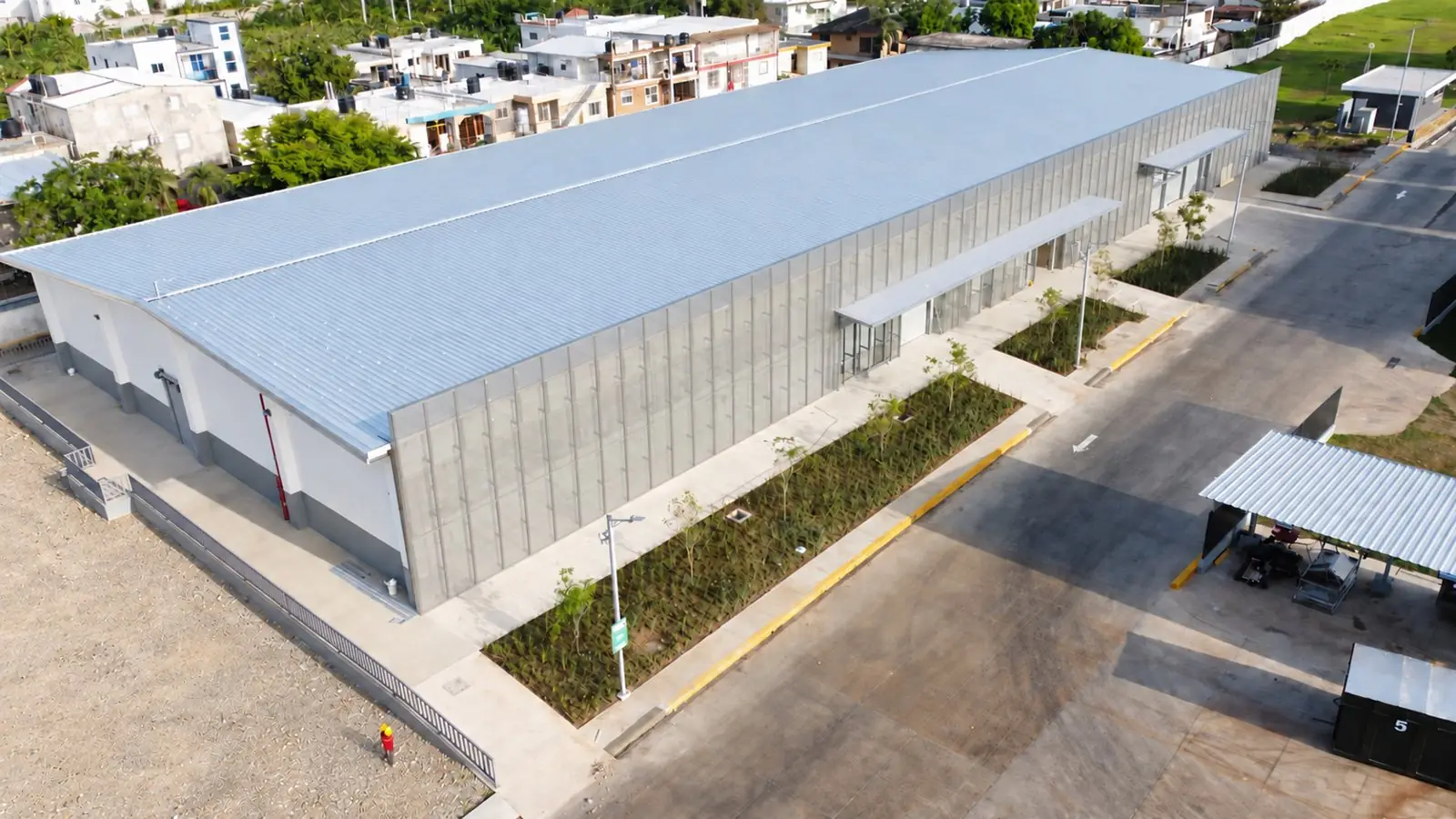 Nave industrial en La Vega, República Dominicana: visualización de acabado sobre la fotografía aérea de la obra, con cubierta metálica uniforme, fachada de paneles perforados completada y entorno sin escombros, tratados digitalmente